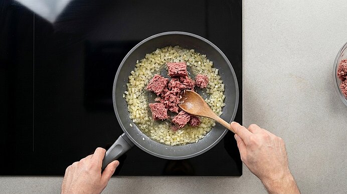 Obenaufnahme einer Pfanne auf dem Herd: glasig gedünstete Zwiebeln, rohes Hackfleisch wird mit einem Holzlöffel eingerührt; Hände halten Pfanne und Löffel.