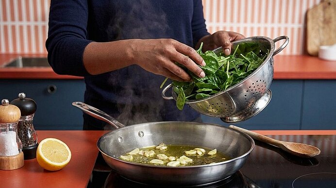 Hände geben frischen Spinat aus einem Sieb in eine Pfanne mit Öl und Knoblauch; Dampf steigt auf. Küchenarbeitsfläche mit Zitrone und Salz‑/Pfeffermühlen im Hintergrund.