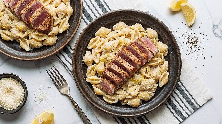 Zwei Teller mit Muschelnudeln in cremiger Sauce, jeweils mit in Scheiben geschnittenem, rosa gebratenem Thunfischfilet garniert.