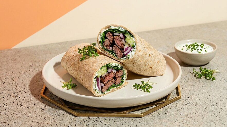 Zwei halbierte Wraps auf einem Teller, gefüllt mit gegrilltem Rindfleisch, Salat, Gurke, roten Zwiebeln und Kräutern; daneben ein Schälchen mit Dip.