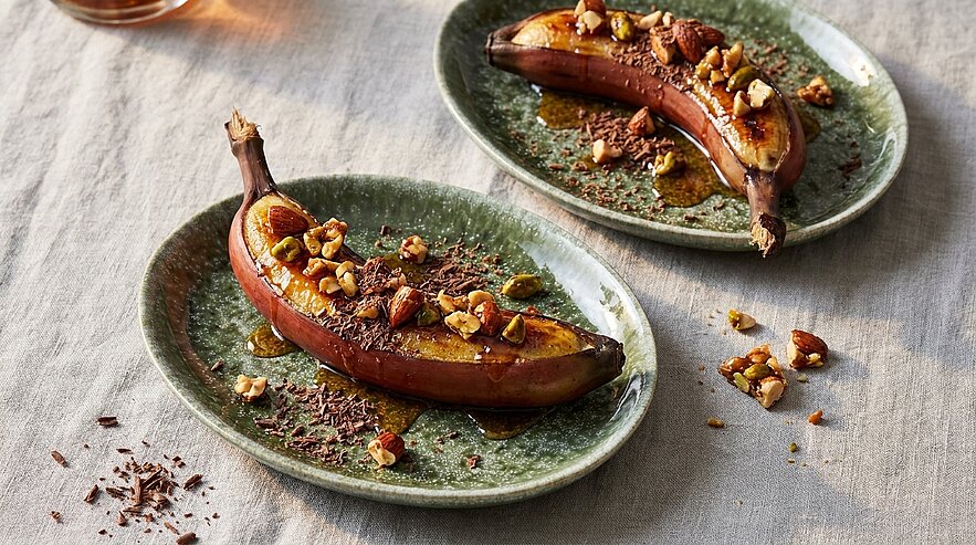 Zwei karamellisierte Bananen in der Schale auf grünen Tellern, beträufelt mit Karamellsauce und garniert mit gehackten Nüssen und Schokoraspeln.
