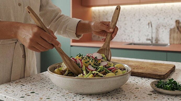 Hände mischen mit zwei Holzlöffeln in einer Keramikschüssel einen frischen Salat mit Radieschenscheiben, Blattsalaten und Kräutern auf einer Küchenarbeitsplatte.