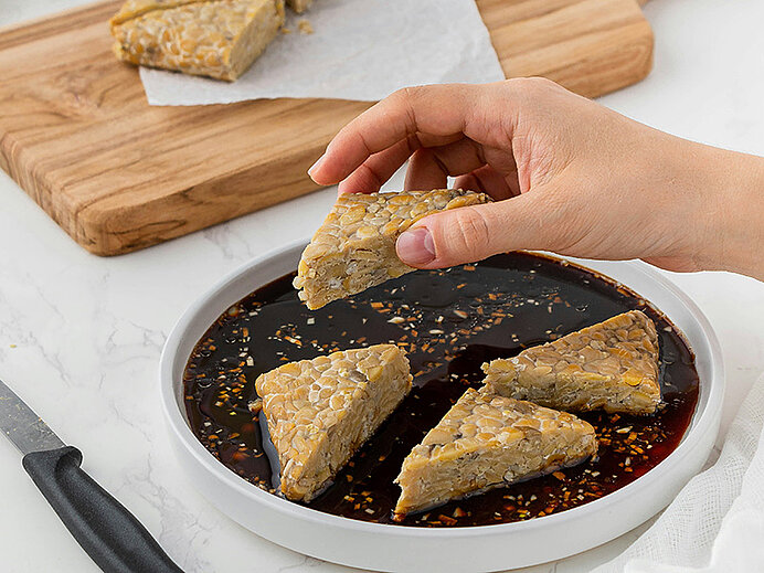 Eine Hand greift nach einem dreieckigen Stück Tempeh, das auf einem Teller mit dunkler Soße liegt. Um das Tempeh sind mehrere gleichmäßig geschnittene Stücke platziert. Im Hintergrund ist ein Holzbrett zu sehen.