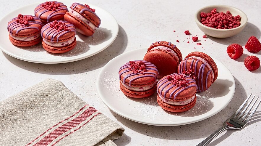 Rosa-rote Himbeer‑Macarons mit violettem Zuckerguss und gefriergetrockneten Himbeerstücken auf weißem Teller; im Hintergrund weiterer Teller, frische Himbeeren und kleine Schale.