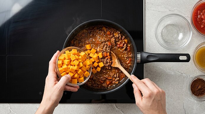 Zwei Hände geben Kürbiswürfel aus einer Glasschale in das köchelnde Chili con Carne im schwarzen Topf.
