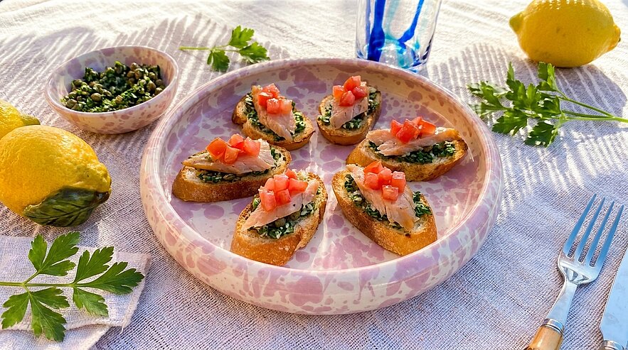 Sechs geröstete Baguettescheiben (Crostini) mit Kräuterpesto und gewürfelten Tomaten als Bruschetta auf einem runden Teller, garniert mit frischer Petersilie.