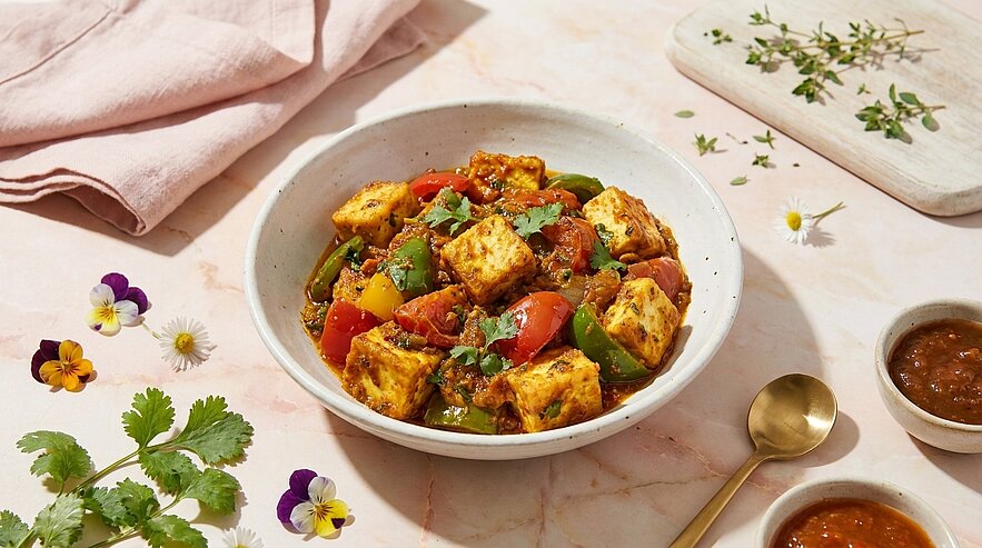 Eine Schüssel mit würzigem Gemüse und Tofu, garniert mit frischem Koriander. Die Mischung enthält bunte Paprika und eine aromatische Soße. Neben der Schüssel liegen zwei kleine Schalen mit Saucen und ein goldener Löffel. Im Hintergrund sind florale Elemente und ein Holzbrett sichtbar.