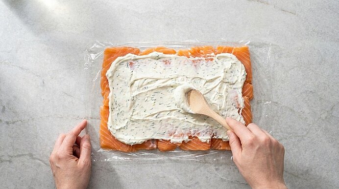 Zwei Hände verstreichen mit einem Holzlöffel weiße Dill-Frischkäsecreme auf ausgelegten Lachsscheiben.