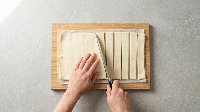 Zwei Hände schneiden ausgerollten Blätterteig auf einem Holzbrett mit einem Messer in schmale Streifen.