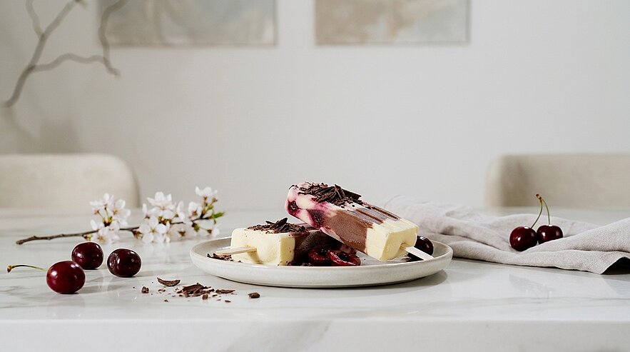 Auf einem Teller liegen aufgeschnittene Scheiben eines Schoko‑Vanille‑Eisdesserts, garniert mit Schokoraspeln und frischen Kirschen; einige Kirschen und Blüten liegen dekorativ daneben.