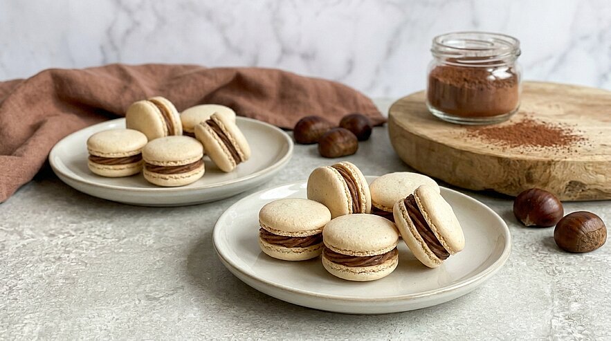 Zwei weiße Teller mit jeweils mehreren hellen Macarons, gefüllt mit dunkler Schokoladencreme, auf hellem Tisch; im Hintergrund Glas mit Kakaopulver und einige Kastanien.