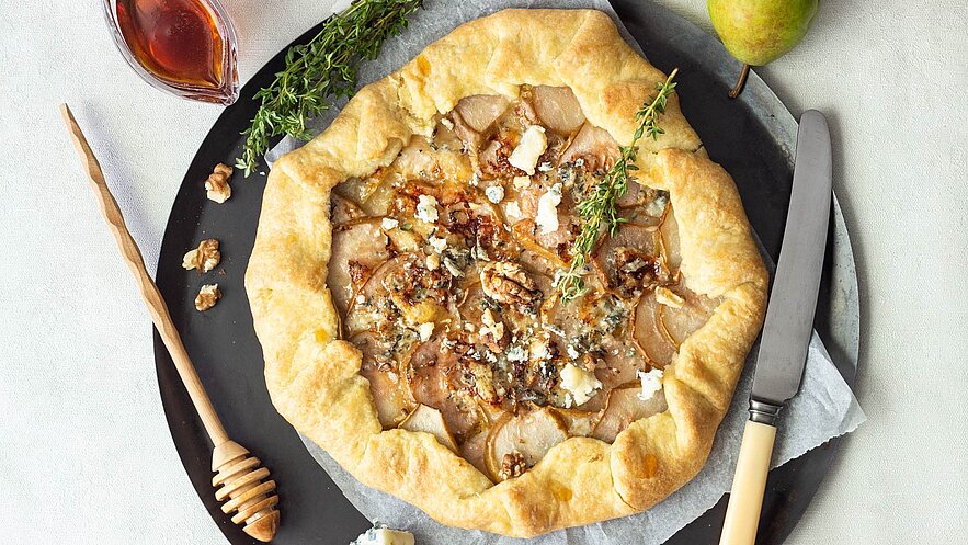 Ein frisch gebackener, rustikaler Kuchen mit einer goldbraunen Kruste, gefüllt mit dünn geschnittenen Birnen, Walnüssen und Käse. Oben sind einige Thymianzweige verteilt. Daneben befindet sich ein Honiglöffel und eine kleine Schale mit Sirup.