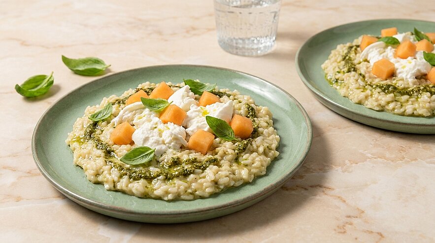 Zwei grüne Teller mit cremigem Risotto, umgeben von grünem Pesto, garniert mit Burrata, Melonenwürfeln und Basilikum.