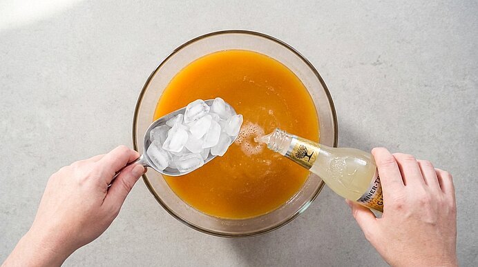 Zwei Hände fügen Eiswürfel und Limonade aus einer Flasche in eine große Glasschüssel mit orangefarbener Bowle.