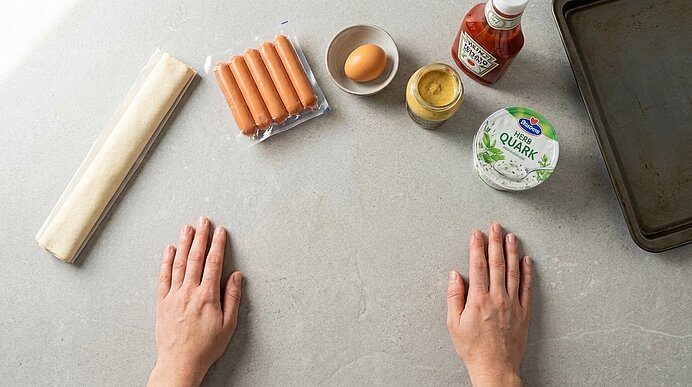 Zwei Hände liegen auf der hellgrauen Arbeitsfläche neben Blätterteig, Würstchen, Ei, Senf, Ketchup und Backblech.