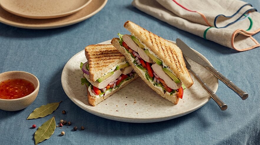 Gegrilltes Toastsandwich, diagonal halbiert und übereinander auf einem Teller, gefüllt mit Salat, Tomate, Avocado und Putenbrust; Messer und Gabel daneben.