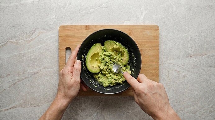 Zwei Hände zerdrücken Avocadofruchtfleisch mit einer Gabel in einer schwarzen Schüssel auf einem Holzbrett.