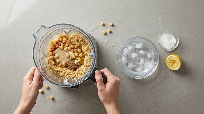 Zwei Hände halten einen Mixer mit Kichererbsen, Tahini und Gewürzen. Daneben stehen Eiswasser, Salz und Zitrone.