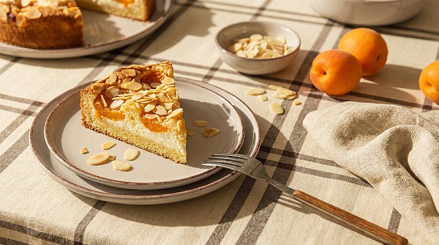Auf einem gedeckten Tisch liegt ein Stück Aprikosenkuchen mit Mandelblättchen auf einem Teller. Daneben befindet sich eine kleine Schüssel mit weiteren Mandeln und frische Aprikosen. Ein Gabel liegt bereit, um den Kuchen zu genießen.