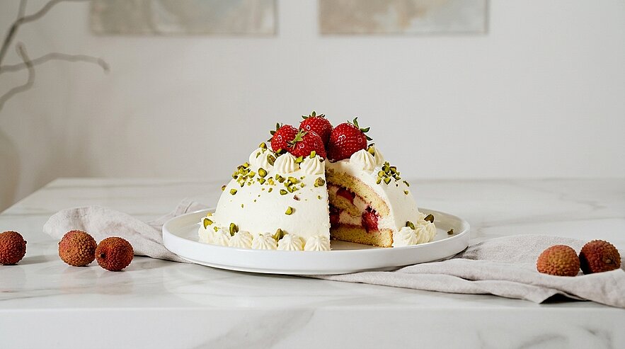 Eine üppige Torte auf einem weißen Teller, dekoriert mit frischen Erdbeeren und Pistazien. Ein Stück der Torte wurde herausgeschnitten, zeigt die bunten Schichten im Inneren. Rund um den Teller liegen einige Litchis auf einem hellen Tisch.