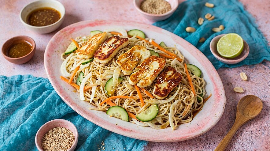 Teller mit Sesam-Nudeln, gemischt mit Gurkenscheiben und Karottenstreifen, belegt mit goldbraun gebratenen Halloumi-Scheiben und Sesam.