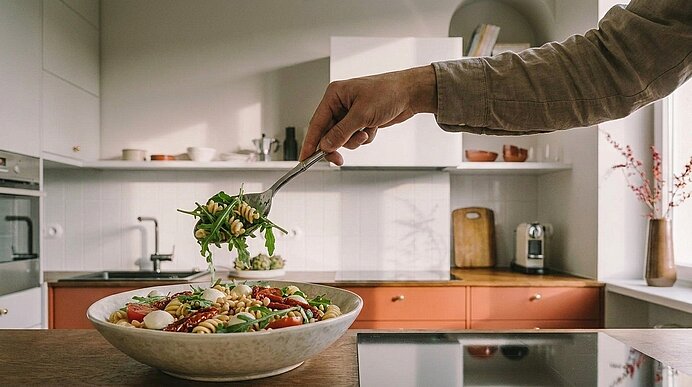 Hand einer Person fügt mit einer Gabel Rucola zu einer großen Schüssel Nudelsalat mit Tomaten, Mozzarella und Kräutern in einer Küchenzeile hinzu.