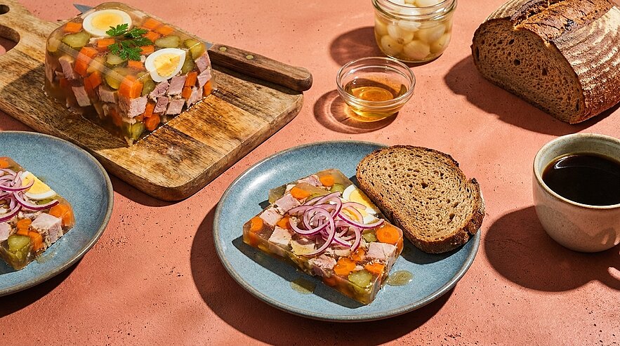Fleischsülze im Aspik auf Tellern, garniert mit eingelegten roten Zwiebeln und Ei, serviert mit dunklem Bauernbrot.