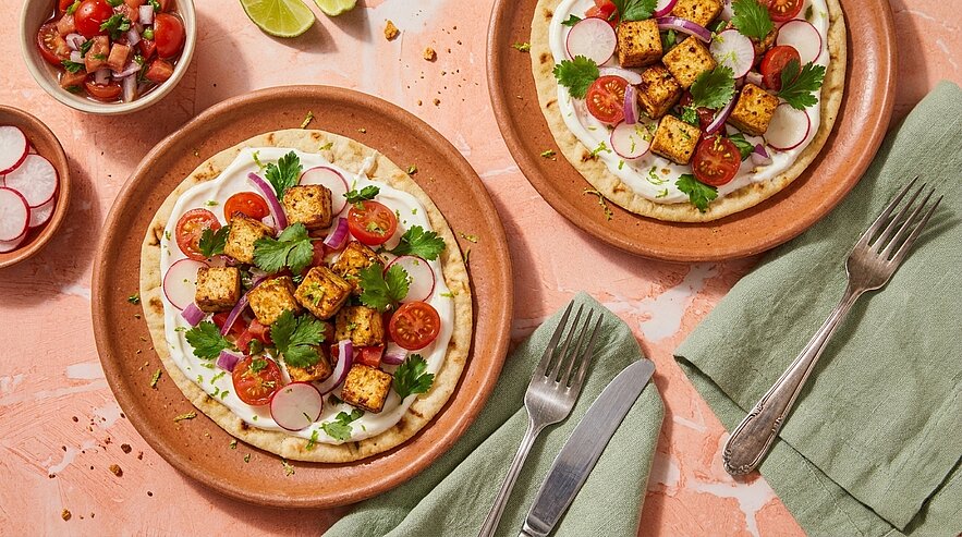Zwei Teller mit Fladenbrot, bestrichen mit Joghurtcreme, belegt mit gegrilltem Tofu, halbierten Kirschtomaten, dünnen Radieschenscheiben und frischem Koriander.