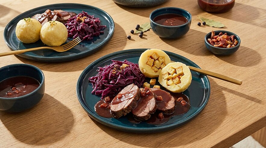 Zwei Teller mit aufgeschnittenem Braten in dunkler Soße, Rotkohl und gefüllten Kartoffelknödeln auf einem Holztisch.