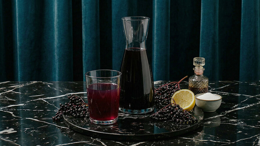 Auf einem dunklen Marmor-Tisch steht eine Glaskaraffe mit dunklem Saft und ein Glas mit der gleichen Flüssigkeit. Umgeben sind sie von Holunderbeeren, einer aufgeschnittenen Zitrone, einer Zuckerschale und einem kleinen Fläschchen. Der Hintergrund ist in einem satten Blau gehalten.