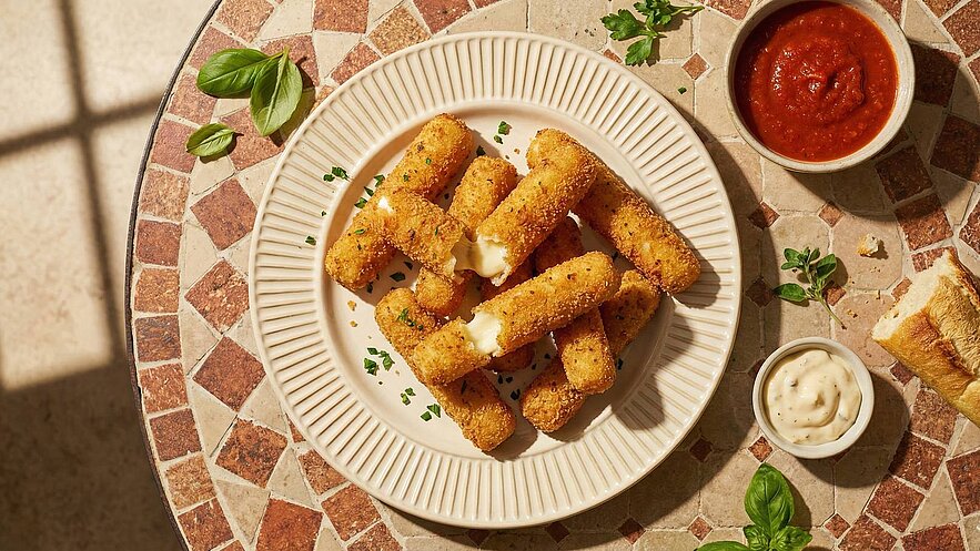 Auf einem runden Teller liegen mehrere knusprige, goldbraune Käse-Sticks, die zum Teil weich und schmelzend sind. Daneben befinden sich zwei Schalen mit Dips: eine rote Tomatensauce und eine cremige weiße Sauce. Der Teller ist auf einem gemusterten Tisch platziert.