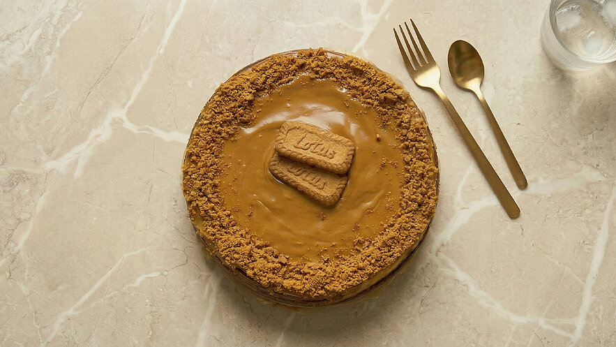 Ein runder Kuchen mit einer glatten, karamellfarbenen Oberfläche, dekoriert mit zerkrümelten Keksen. Oben liegen zwei Keksstücke mit der Aufschrift "Lotus". Der Kuchen steht auf einer eleganten, hellen Tischplatte, daneben liegen eine Gabel und ein Messer.
