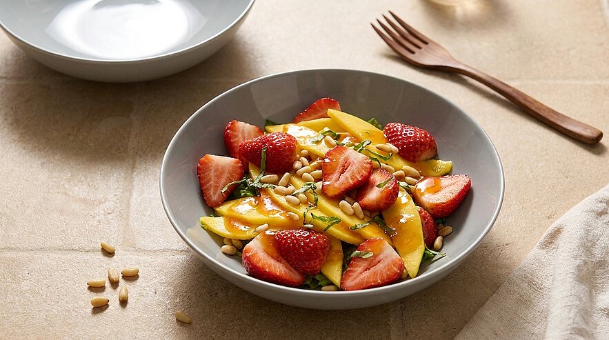 Eine Schüssel mit frischem Obstsalat, bestehend aus geschnittenen Mangostücken und Erdbeeren, garniert mit Pinienkernen und frischen Kräutern. Die Schüssel ist auf einer neutralen Oberfläche platziert, daneben liegt ein Holzlöffel.