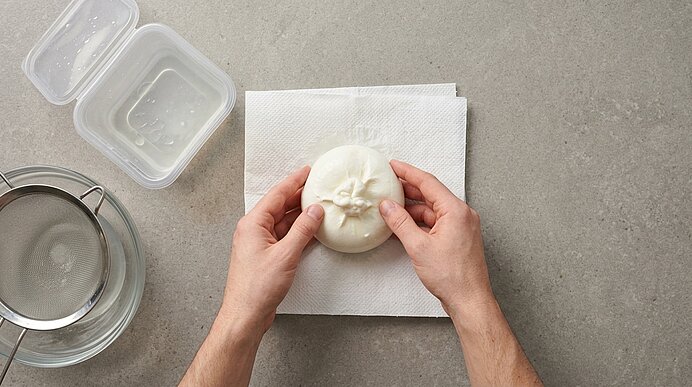 Zwei Hände halten eine frische Burrata zum Abtropfen auf Küchenpapier auf einer hellgrauen Steinarbeitsplatte.