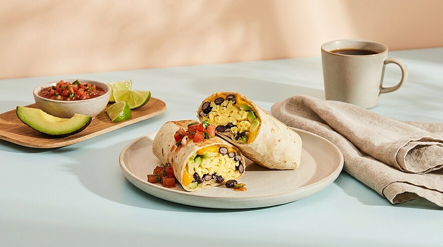 Zwei halbierte Frühstücks-Burritos auf einem Teller, gefüllt mit Rührei, schwarzen Bohnen, Mais und Käse; daneben Schale mit Salsa, Avocado und Limette sowie eine Tasse Kaffee.
