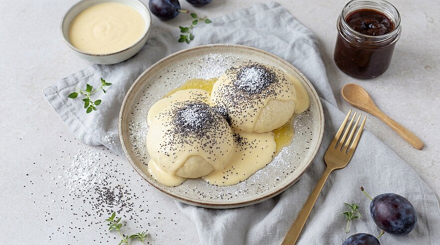 Zwei gefüllte Germknödel auf einem Teller, übergossen mit Vanillesoße und bestreut mit Mohn und Puderzucker.