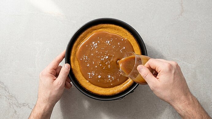 Zwei Hände gießen flüssiges Salzkaramell aus einem Glaskännchen auf den gebackenen Kürbis-Cheesecake in einer Springform.