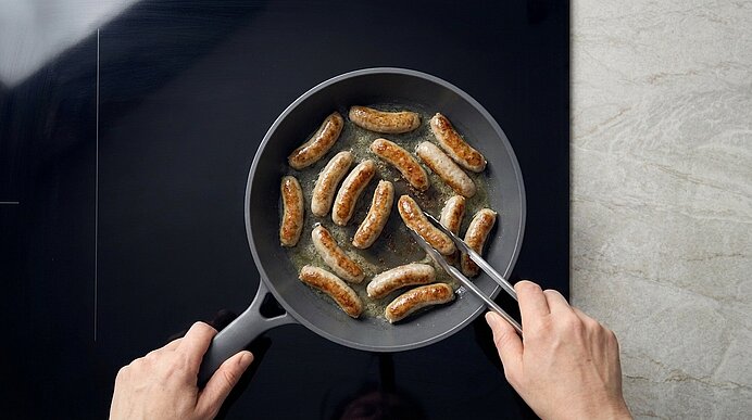 Obenansicht: Hände braten mehrere kleine Bratwürste in einer Pfanne; eine Hand hält den Griff, die andere wendet die Würste mit einer Zange.