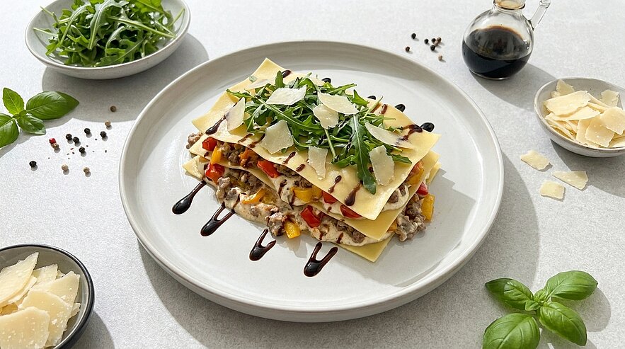 Mehrschichtige Gemüse-Lasagne, gefüllt mit Pilzen und Paprika, garniert mit Rucola, gehobelten Parmesanspänen und Balsamico-Reduktion auf einem weißen Teller.