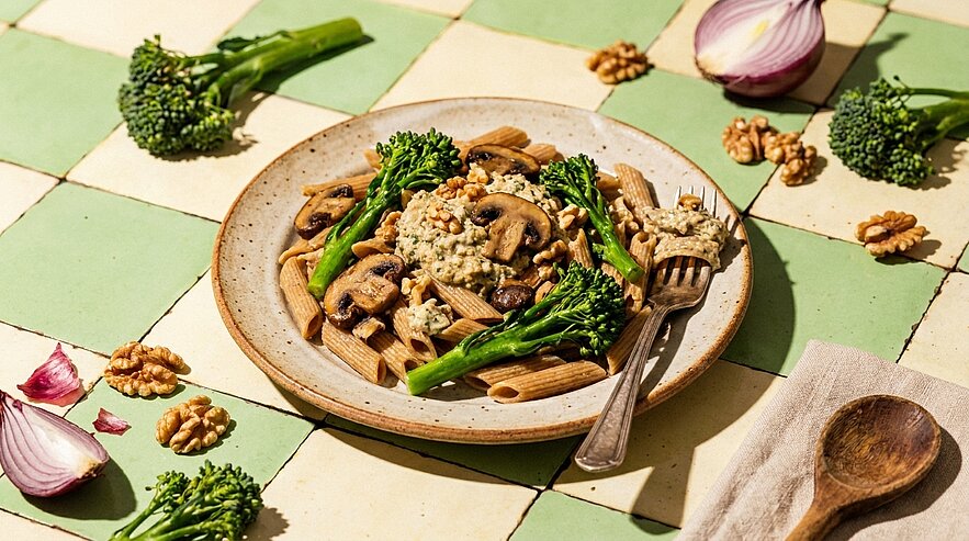 Teller mit Vollkorn-Penne, sautierten Champignons, Broccolini und zerbröselten Walnüssen, eine Gabel liegt am Rand.