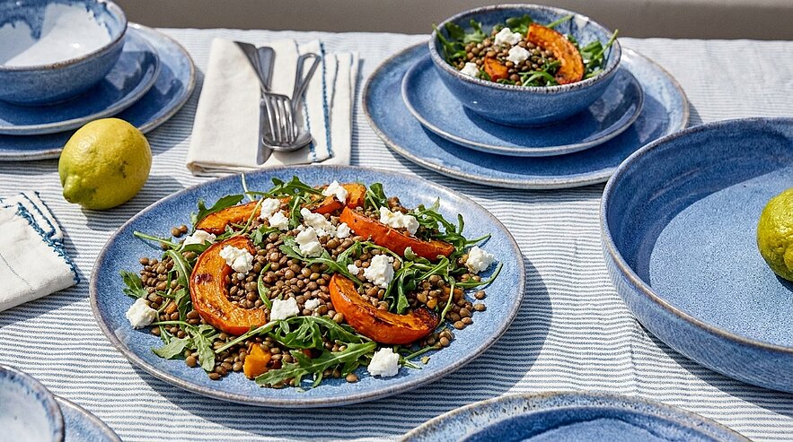 Blauer Keramikteller mit Linsensalat aus Beluga-Linsen, Rucola, gerösteten Kürbisspalten und zerbröseltem Feta, angerichtet auf einem hellen Tisch.