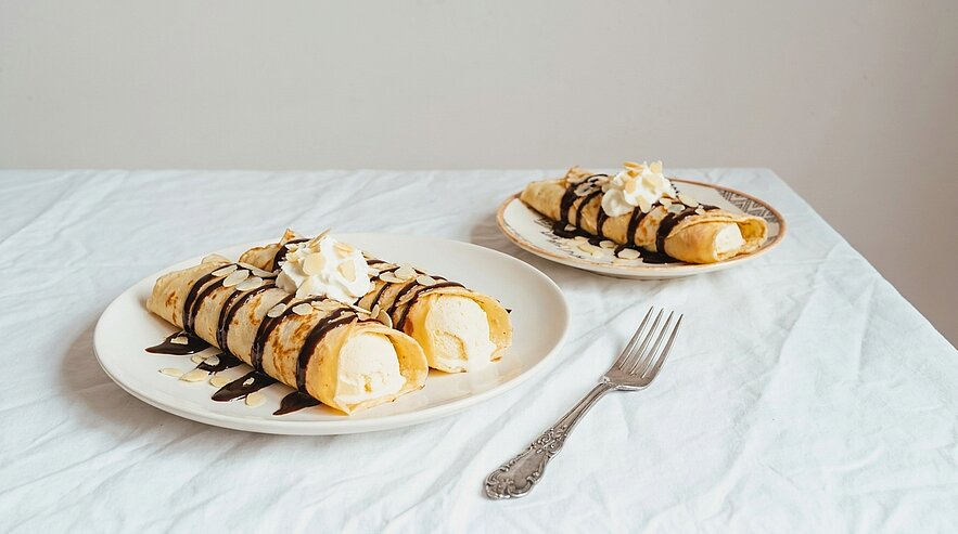 Zwei Teller mit gefüllten Crepes, jeweils mit Vanilleeis, Schokoladensoße und Schlagcreme garniert; eine Gabel liegt neben einem Teller.