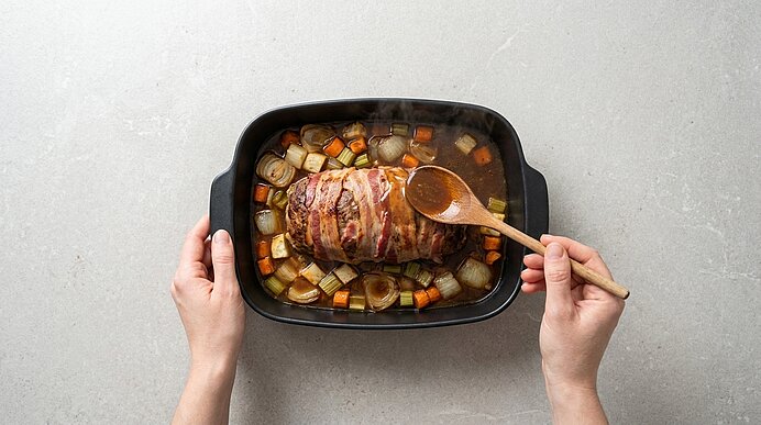 Hände halten einen Bräter mit einem im Speckmantel gebratenen Braten, umgeben von Zwiebeln, Karotten und Kartoffeln; der Braten wird mit einem Holzlöffel bestrichen.