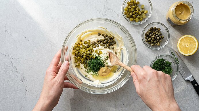 Zwei Hände verrühren Mayonnaise, Cornichons, Kapern, Dill und Senf mit einem Holzlöffel in einer Glasschüssel.