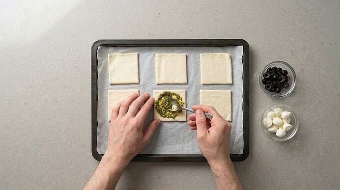 Zwei Hände verteilen mit einem Löffel grünes Pesto auf Blätterteigquadraten auf einem Backblech.