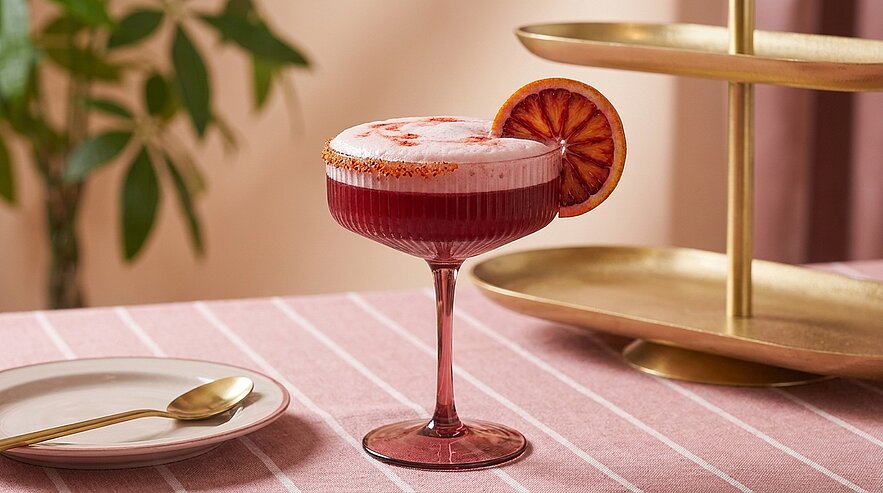 Roter Schaumcocktail in einem rosa Coupéglas, garniert mit einer halben Blutorangenscheibe.