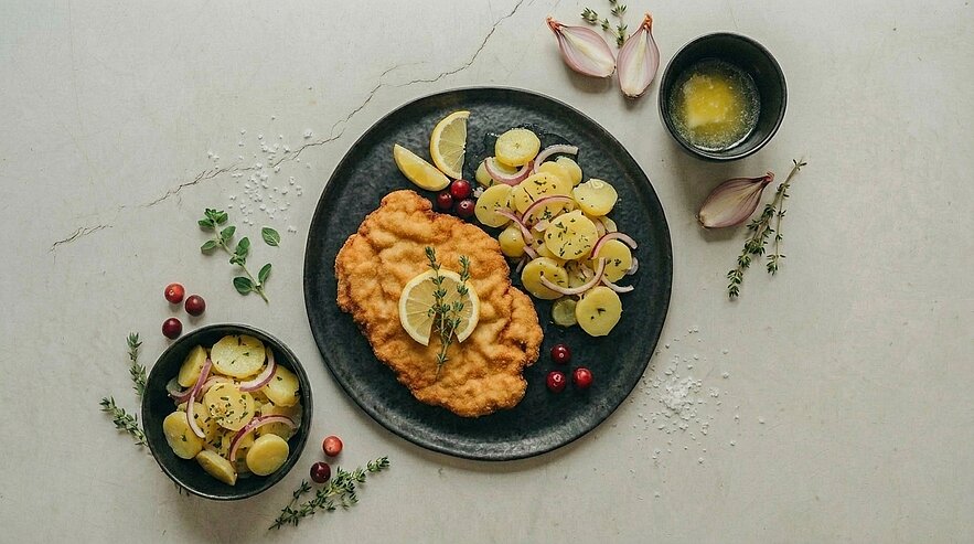 Paniertes Schnitzel mit Zitronenscheiben auf einem Teller, dazu Kartoffelsalat mit dünnen Kartoffelscheiben, roten Zwiebeln und frischen Kräutern.