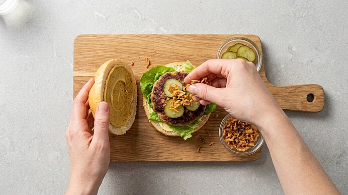 Zwei Hände streuen Röstzwiebeln auf eine Frikadelle mit Senfgurken im Brötchen auf einem Holzbrett.
