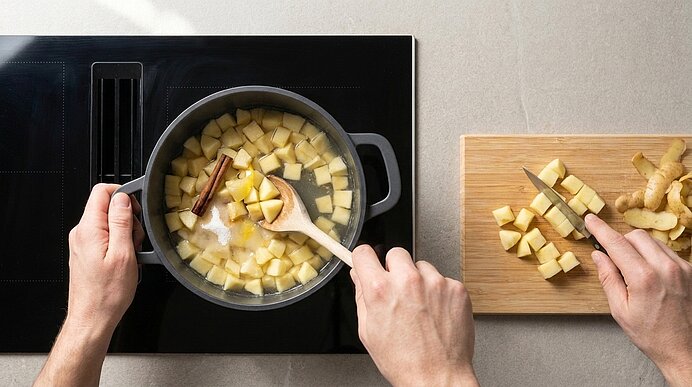 Obere Draufsicht auf Hände, die gewürfelte Äpfel mit Zimtstange und Zucker in einem Kochtopf rühren, während die andere Hand auf einem Schneidebrett weitere gewürfelte Äpfel und frischen Ingwer mit einem Messer schneidet.