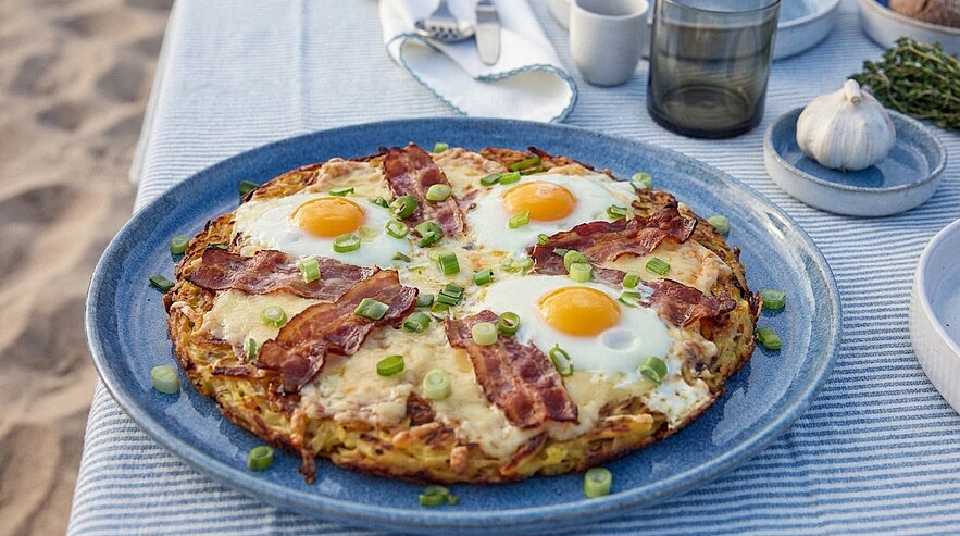 Großer knuspriger Kartoffelrösti auf Teller, belegt mit vier Spiegeleiern, knusprigem Speck, geschmolzenem Käse und gehackten Frühlingszwiebeln.